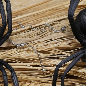 May include: Two black spider decorations made of woven straw, with silver chain accents, on a background of dried wheat stalks.