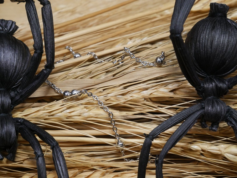 May include: Two black spider decorations made of woven straw, with silver chain accents, on a background of dried wheat stalks.