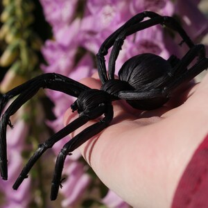 May include: A black spider made of woven material, held in a person's hand. The spider has eight legs and a round body.