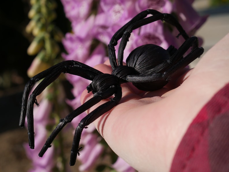 May include: A black spider made of woven material, held in a person's hand. The spider has eight legs and a round body.