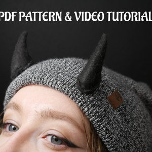 Puede incluir: Un gorro gris con dos cuernos de fieltro negro unidos a la parte superior. El gorro tiene una pequeña etiqueta marrón en el lateral.