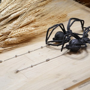 May include: Two black spider earrings with silver chains and beads. The spiders are made of woven material and have a realistic appearance.