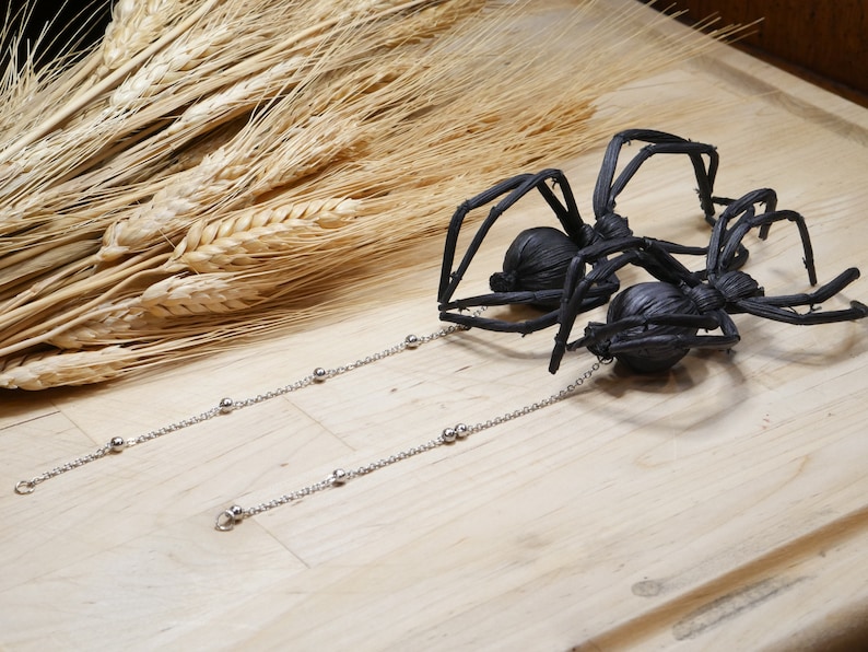 May include: Two black spider earrings with silver chains and beads. The spiders are made of woven material and have a realistic appearance.