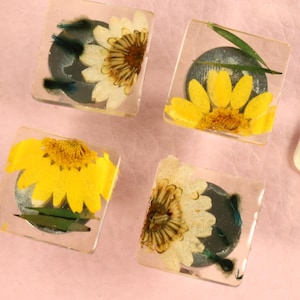 Puede incluir: Cuatro pisapapeles cuadrados de resina con flores prensadas. Cada pisapapeles presenta un diseño de flor diferente con pétalos amarillos y un centro oscuro.