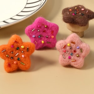 Könnte beinhalten: Fünf filzförmige Sterne mit bunten Streuseln. Die Sterne sind in den Farben Rosa, Orange, Blau, Braun und Türkis.