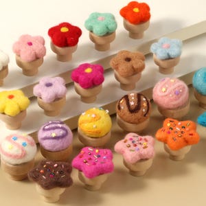 May include: A collection of 20 felt cupcake toppers in various colours and designs, including flowers, stars, and swirls. The toppers are attached to wooden bases.
