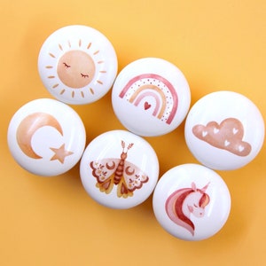 Op de afbeelding: Set van zes witte keramische ladegrepen met verschillende pastelkleurige illustraties. De ontwerpen omvatten een zon, een maan, een regenboog, een wolk, een vlinder en een eenhoorn.