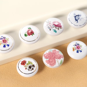 Könnte beinhalten: Satz mit sechs weißen Keramik-Schrankknöpfen mit goldener Einfassung und Blumenmustern. Jeder Knopf zeigt ein anderes Blumenmuster in den Farben Rosa, Blau, Gelb und Grün.