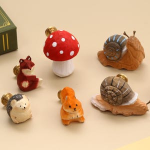 May include: A collection of seven carved wooden drawer knobs featuring animal and nature shapes, including a red and white mushroom, two snails with brown shells, a red fox, a brown hedgehog, an orange squirrel, and a light pink rabbit figure.