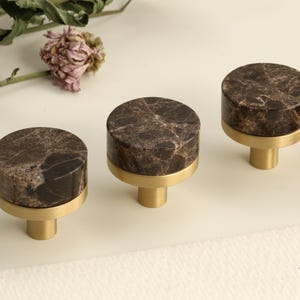 May include: Four cabinet knobs with brown marble tops and gold-coloured brass bases. The cylindrical knobs are displayed on a light surface, with dried flowers in the background. Each knob is approximately 3 cm in diameter.