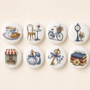 May include: A set of eight round ceramic cabinet knobs, each featuring a distinct illustration including a teacup with a croissant, a bistro chair and table, a bicycle with flowers, a clock, a window with an awning, a perfume bottle, a key, and books.
