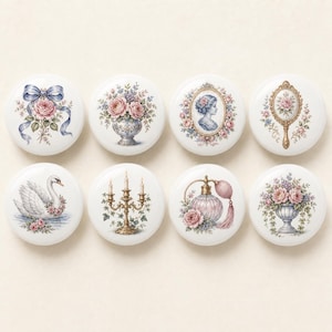 May include: A set of eight round white ceramic knobs featuring colorful illustrations of a blue ribbon with roses, floral vases, a cameo portrait, a hand mirror, a white swan, a gold candelabra, a perfume bottle, and a floral arrangement.