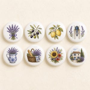 May include: A set of eight round white ceramic knobs featuring colorful illustrations including lavender bouquets, olive branches, lemons, a cicada, a blue and white pitcher, a bread basket, sunflowers with poppies, and a stone house with lavender.