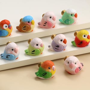 May include: A collection of ten colorful, hand-painted bird figurines. Each bird has a unique color scheme, including blue, orange, yellow, green, and pink. The birds are perched on a white surface, showcasing their detailed craftsmanship.