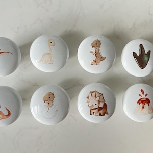 Könnte beinhalten: Acht weiße Keramik-Schrankknöpfe mit Dinosaurier-Illustrationen. Die Knöpfe zeigen verschiedene Dinosaurier in Braun-, Orange- und Grüntönen, darunter ein Pterodactylus, ein Brontosaurus und ein Triceratops. Ein Knopf hat einen Dinosaurierfußabdruck.