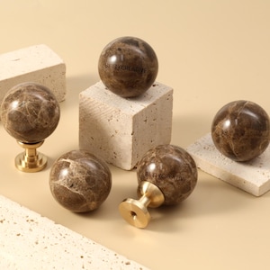 Könnte beinhalten: Braune Schrankknöpfe aus Marmor mit Messingbasen werden auf einer beigefarbenen Oberfläche präsentiert. Einige Knöpfe befinden sich auf kleinen, hellfarbigen Steinblöcken. Die Knöpfe haben eine polierte Oberfläche und ein einzigartiges Aderungsmuster.