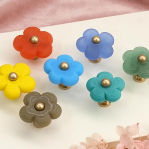 Puede incluir: Una colección de siete pomos para armarios con forma de flor en varios colores, incluyendo rojo, amarillo, azul, azul claro, verde, verde azulado y gris. Cada pomo presenta un acabado mate y una pieza central metálica dorada.