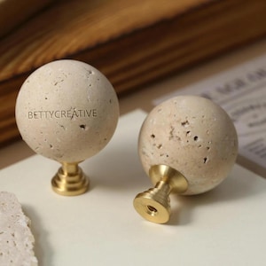Peut inclure: Deux boutons de meuble décoratifs. Chaque bouton présente un dessus en pierre sphérique beige avec une surface texturée. La base est en métal doré. Le texte "BETTYCREATIVE" est imprimé sur l'un des boutons.