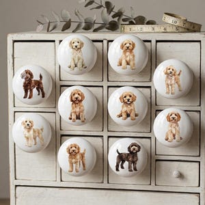 May include: A collection of white ceramic cabinet knobs, each featuring a different adorable puppy illustration. The puppies are depicted in various poses and colors, including golden, brown, and black. The knobs are arranged on a white cabinet.