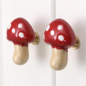 May include: Two mushroom-shaped cabinet knobs featuring a glossy red cap with white spots and a smooth beige stem. Each knob is attached to a circular brass-toned metal base, providing a functional handle for furniture doors or drawers.