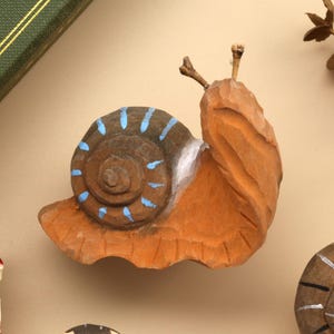 May include: A hand-carved wooden snail figurine featuring a light brown body and a dark brown shell with painted light blue stripes. The snail has two small, thin antennae extending from its head, and the wood shows visible carving marks throughout.