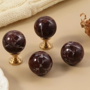 Pode incluir: Quatro puxadores decorativos para armários com aparência de mármore vermelho escuro e veios brancos. Os puxadores são esféricos e montados em bases douradas. Os puxadores estão dispostos sobre uma superfície bege clara.
