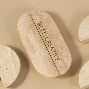 Puede incluir: Colección de elementos de piedra beige claro, que incluyen una pieza ovalada con el texto "BETTYCREATIVE" y una línea vertical, y varias piezas semicirculares. Los elementos están dispuestos sobre una superficie marrón claro.