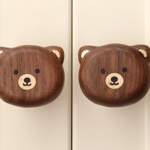 May include: Two dark brown wooden cabinet knobs shaped like bear heads. Each knob features a smooth, rounded design with carved ears, two small black circular eyes, and a lighter-colored muzzle area with a black nose and a small curved mouth.