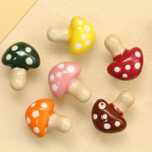 Pode incluir: Uma coleção de seis pequenos puxadores em forma de cogumelo em várias cores, incluindo verde-escuro, amarelo, vermelho, rosa, laranja e castanho. Cada puxador apresenta um acabamento liso e brilhante com pintas brancas no chapéu.