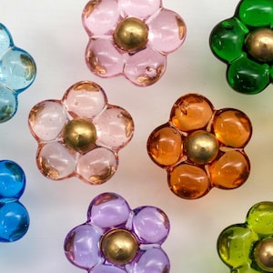 Peut inclure: Un ensemble de huit boutons de placard en forme de fleur en verre coloré translucide. Chaque bouton présente cinq pétales arrondis dans des tons bleu, rose, vert, orange, jaune et violet, avec un dôme central en métal poli couleur laiton.
