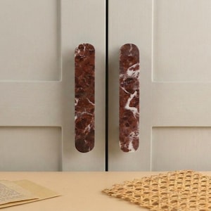 May include: Two vertical, elongated cabinet handles made of deep red marble with prominent white veining. The handles have rounded top and bottom edges and are mounted onto a light-coloured, panelled cabinet door surface with a smooth, matte finish.