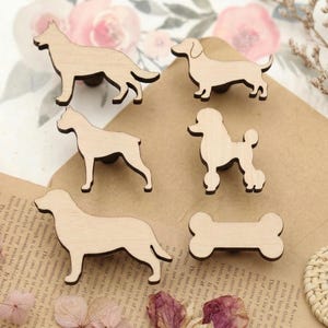 May include: A selection of wooden dog-shaped cutouts, featuring a German Shepherd, Dachshund, Poodle, and a bone. The light brown cutouts have a natural wood grain. They are displayed on a textured surface with floral decorations.