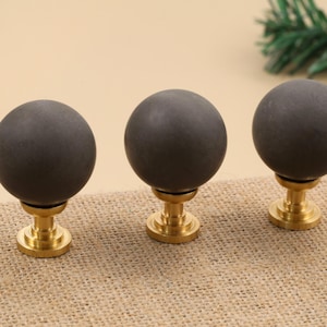 May include: Three dark gray spherical cabinet knobs with a brass base are arranged on a tan surface. The knobs have a matte finish and a simple, modern design. The brass bases add a touch of elegance.