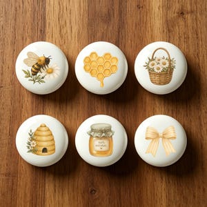May include: Set of six white ceramic cabinet knobs with bee, honeycomb, basket of flowers, beehive, jar of honey, and bow designs. Each knob features a different illustration in shades of yellow, brown, and green, perfect for a kitchen or craft room.