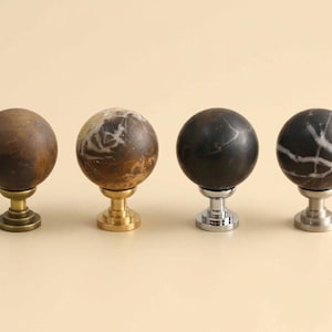 May include: Four spherical cabinet knobs arranged in a row. The first is brown stone with an antique brass base, the second is mottled brown and white stone with a gold base, the third is black stone with a chrome base, and the fourth is black and white veined stone with a brushed nickel base.
