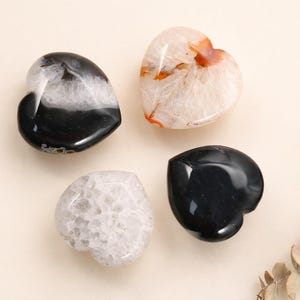 Puede incluir: Cuatro piedras en forma de corazón en tonos negro, blanco y naranja. Las piedras tienen una textura pulida y suave. Una piedra es negra, otra es blanca con vetas negras, y las otras dos son blancas y naranjas. Una ramita de hojas secas en el fondo.