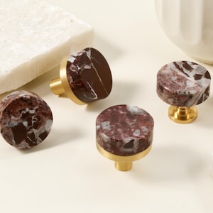 Op de afbeelding: Vier decoratieve kastknoppen met een marmerontwerp. De knoppen hebben een mix van bordeauxrode en witte kleuren. De bases zijn goudkleurig. De knoppen zijn gemaakt van steen en metaal. De knoppen zijn voor meubels.