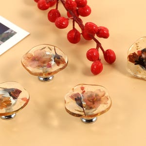 Puede incluir: Cuatro pomos decorativos para armario con un diseño floral encapsulado en resina transparente. Los pomos son de color marrón claro con una base plateada.