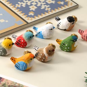 Puede incluir: Una colección de ocho figuritas de pájaros de madera talladas a mano en varios colores. Cada pájaro tiene un pomo dorado en la parte superior. Los pájaros están pintados en tonos de amarillo, azul, rojo, verde, marrón y rosa, con detalles en blanco y negro.