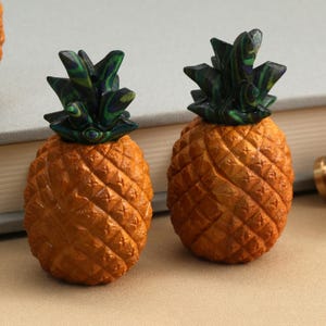 May include: Two pineapple-shaped cabinet knobs featuring textured, golden-brown bodies and dark green, marbled-patterned leaves. Each knob has a metallic base attachment, showcasing a detailed, realistic fruit design for furniture hardware.