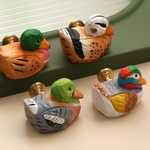 May include: Four decorative duck-shaped cabinet knobs. Each knob is hand-carved and painted with vibrant colours. The ducks have green, orange, white, and blue heads, with detailed wings and bodies. The knobs are mounted on gold-coloured hardware.