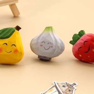 Peut inclure: Trois figurines en bois sur le thème des aliments : une poire jaune avec une feuille verte, un bulbe d'ail blanc et une fraise rouge avec un dessus vert. Chacun a un visage souriant et des joues roses. Les figurines sont sur une surface brun clair.
