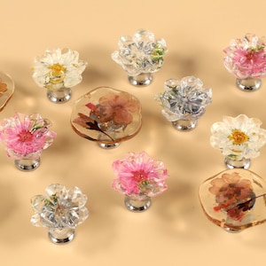 Op de afbeelding: Assortiment van heldere glazen kastknoppen met geperste bloemen erin. De knoppen zijn in verschillende vormen en hebben verschillende bloemarrangementen.