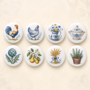 May include: A set of eight round white ceramic knobs featuring colourful illustrations of a rooster, a hen, a blue and white pitcher with flowers, a lidded tureen, an artichoke, a pear branch, a wheat sheaf, and a potted herb plant.