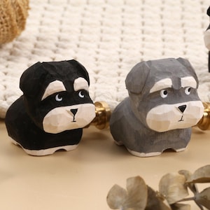 Op de afbeelding: Houten hondenbeeldjes in zwart en grijs, met lichtgekleurde gezichten en goudkleurige hardware. De hondenbeeldjes zijn gesneden met gedetailleerde kenmerken en staan op een lichtgekleurd oppervlak. De achtergrond bevat een geweven textuur en een houten blok.