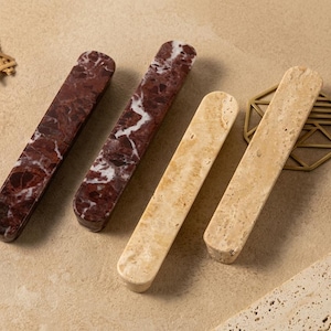 May include: Four rectangular cabinet pulls with rounded ends. Two pulls are made of dark burgundy stone with white veining, and two pulls are made of light beige travertine stone with a porous, textured surface and natural colour variations.