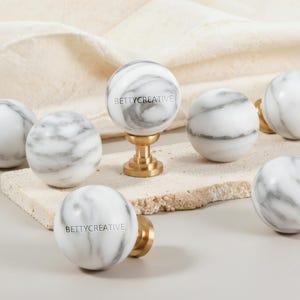 May include: A collection of marble sphere cabinet knobs with gray veining. Some knobs have brass hardware. The text "BETTYCREATIVE" is printed on some of the knobs. The knobs are displayed on a light-colored surface.