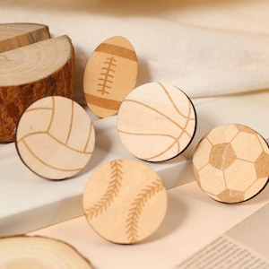 May include: Five wooden ornaments shaped like different sports balls. The ornaments are in the shape of a baseball, basketball, football, soccer ball, and volleyball. The ornaments are all made of light brown wood and have a natural finish.