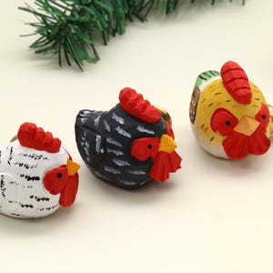 Puede incluir: Tres tiradores decorativos con forma de gallina. Uno es blanco con detalles negros, otro es negro con detalles blancos y el tercero es amarillo, rojo y verde. Cada uno tiene una cresta y una barbilla rojas. Los tiradores son de madera.