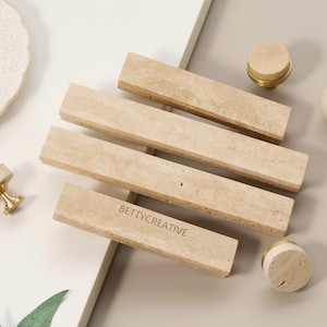 Może przedstawiać: Zestaw beżowych elementów kamiennych, w tym prostokątne uchwyty i okrągłe gałki z mosiężnymi akcentami. Na jednym uchwycie widnieje napis "BETTYCREATIVE". Elementy są ułożone na białej i beżowej powierzchni.